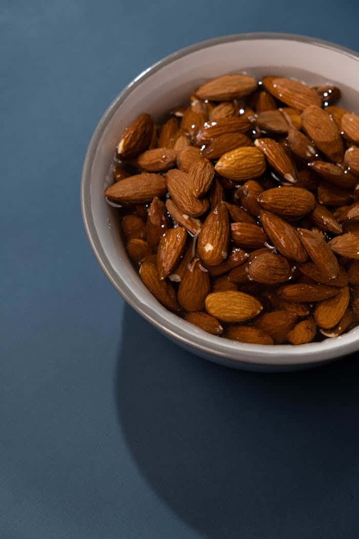Almonds soaking in water in a white bowl on a blue background, promoting healthy snacking and nutrition.